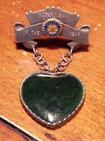Close-up front view of the sterling silver brooch showing the Rotary International emblem, engraved banner, and heart-shaped jade pendant attached with two chains.