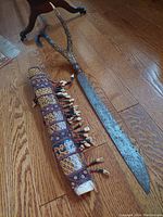 Full view of the machete with sheath on wooden floor showing the blade, handle, and decorated sheath with bead adornments.