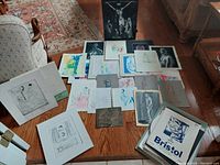 Overview of vintage portfolio artworks by Danielle Sleasman, showing various loose sheets of art including sketches, drawings, and watercolor paintings spread on a table with Bristol board folder visible.