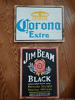 Photo of both wooden signs showing Corona Extra and Jim Beam Black designs, arranged on wooden floor.