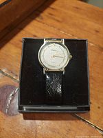 Front view of the vintage Orleans Quartz wristwatch in a black box, showing the round gold-tone case, light grey dial, and textured black leather strap.