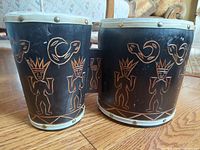 Close-up photo showing the engraved tiki tribal design on the wooden body of the two drums in black with tan accents.