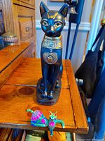Photo showing full view of the black Egyptian cat statue with gold detailing and the colorful small wooden cat figurine on wooden furniture.
