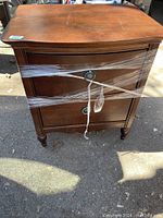 Front view of the wooden 2 drawer end table with plastic wrap securing it, showing the drawer handles and overall shape.