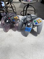 Two Nintendo 64 controllers, one grey and one translucent purple, with attached cables and proprietary connectors, shown from the front.