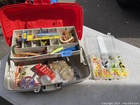 Red plastic fishing tackle box with various fishing lures and accessories inside along with a clear tackle box organizer with soft plastic lures beside it.