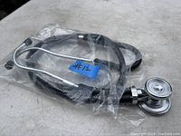 Black stethoscope device with chrome metal chest piece and ear pieces lying coiled on a gray surface, partially wrapped in clear plastic bag with blue label #12 attached.