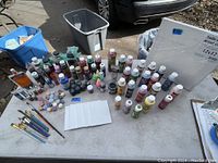 Wide view of the lot showing multiple bottles of acrylic paint, brushes arranged on table, small paint jars and tubes, and wrapped canvases.