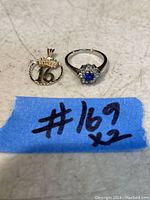 Photo of a 10K gold ring with a blue stone surrounded by small clear stones and a 10K gold pendant featuring a crown and number 16.