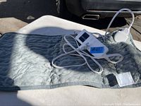 Gray rectangular heating pad with attached temperature controller and power plug, displayed outdoors on a surface with a car in the background.