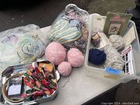Photo showing various yarn balls, skeins of embroidery floss, crochet hooks, sewing tools and craft kits arranged on a surface.