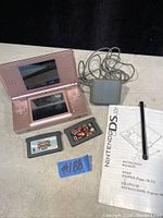 Pink Nintendo DS Lite handheld console open with dual screens visible, two Game Boy Advance cartridges in front, charger cable attached, instruction manual, and black stylus pen.