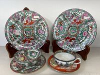Two vintage Chinese Famille Rose Medallion porcelain plates on wooden stands with matching bowl and a separate small tea cup and saucer in front, all showing detailed hand-painted traditional Chinese scenes and floral designs, with gold accents.