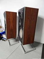 Two B&O Beovox S75 speakers without grille covers showing driver layout and walnut veneer cabinets resting on metal stands