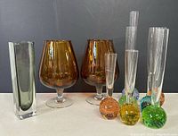 Photo showing all 9 items: 7 bubble bud vases mostly with colored bubble bottoms in green, yellow, orange, plus a tall rectangular smoky gray vase and 2 amber brandy glasses.