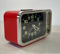 Left angled view showing red casing, clock face with luminous numbers, hands, chrome bezel and alarm controls