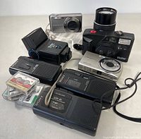 Lot contents arranged showing two Sony mini cassette recorders, several mini cassette tapes, a silver compact digital camera, a Vivitar Optio ECO digital camera, and a Vivitar film camera with zoom lens.