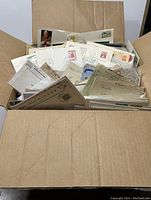 Cardboard box filled with numerous first day covers, covers, and postal items seen inside, showing the variety of stamps and postmarks.