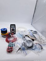 Wide shot showing digital multimeter, spools of wire, multiple cables, and test leads packed in plastic bags, all arranged on white surface.