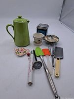 Full lot showing all assorted kitchen items including green teapot, small floral dish, plant pot, spatulas, grater, spoons, thermometer, and tin container.