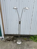 Full view of the silver-tone metal double gooseneck standing floor lamp with two dome-shaped shades and weighted round base under natural light against a white corrugated backdrop.