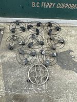 Nine circular metal plant stands with three small wheels each, arranged on concrete. Black with signs of age and patina.