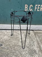 Full view of black wrought iron jardiniere stand showing circular top and angled legs with curled feet, with diamond-shaped accents on the frame.
