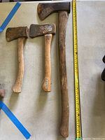 Three axes laid out side by side: one large axe with long handle and two smaller axes with shorter handles, placed next to measuring tape for scale.