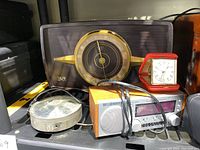 Overview of vintage clocks and clock radios including a large mid-century style analog clock radio with prominent circular face, a small red and white travel clock, and various other clock radios and clocks in wood, metal, and plastic finishes.