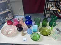 Overview of all colored glass items on white table showing variety of bowls, vases, and pitchers in multiple colors.