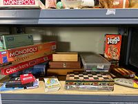 Shelf view showing boxed Monopoly, Checkers, Jenga games along with wooden boxes, stacked playing cards, and other game accessories.