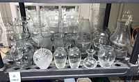 Many glass vases of varying shapes and sizes, clear glass, arranged on a shelf, some vintage style, some etched and textured.