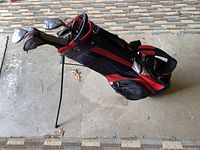 Black and red golf bag with multiple golf clubs inside placed on concrete floor showing stand extended