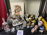Photo of various porcelain and ceramic figurines, decorative bells, glass candle holders, shoe-shaped planters, and a framed cross stitch artwork.