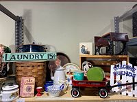 Overview of enamel pot, cup, teapot, trays, small red Radio Flyer wagon with accessories, wooden doll desk with chair, boxes, woven basket, and decorative signs on shelving.