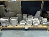 Shelf containing multiple stacks of assorted plates, bowls, ramekins, and small loaf pans, all white and needing cleaning.