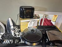 Wide shot of two toasters, two irons, Elite Gourmet round electric griddle with glass lid, and Mickey Mouse waffle maker on a countertop. Items are plugged in or have cords visible and appear used needing cleaning.