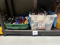 Green plastic tray and clear HDX bin containing assorted metal and wooden utensils including pizza cutter, tongs, spatulas, whisks, strainers, can opener, measuring spoons, silverware and rolling pin