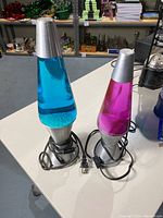 Photo showing two lava lamps with silver bases and caps, one with blue liquid and one with pink liquid, both plugged in with cords wrapped at bases.