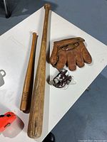 Two wooden baseball bats, one longer and one shorter. Vintage brown leather baseball glove. Pair of whistles tied together with a cord.