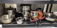 Shelf display of various stainless steel cookware including pots, skillets, double boiler, some with lids and a red non-stick skillet pan.