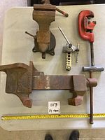 Top-down view of two heavy duty cast iron vices, red pipe cutter, pipe threader, and pipe cutting accessory on table with ruler for scale.