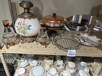 Vintage lamp with floral glass globe and ornate metal base, copper fondue pot with lid and burner stand, two glass coffee carafes with copper lids and warmers, Royal Albert serving plate and casserole carrier on metal shelving rack.