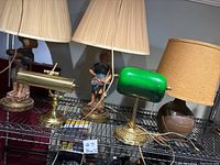Five vintage lamps arranged on wire shelving including figurine boy and girl lamps, green bankers lamp, brass piano lamp, and brown crockery lamp.