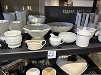 Image showing assorted white milk glass and other white glass pieces on a black shelf including footed bowls, mugs, a creamer, and a lidded jar