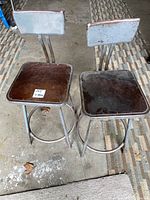 Top-front angle showing both vintage industrial stools side by side, highlighting metal backrests and wooden square seats with wear.
