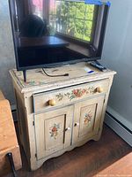 Front view of vintage wooden cabinet with floral hand painting, showing drawer and doors, surface wear evident.