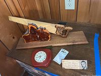 Two wooden clocks, one shaped like Kentucky and one red plastic framed clock, with a vintage student name plate and a large wooden saw behind them.