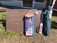 Overall view of brown folding table, badminton set box, Wilson tennis racket, and folded item in blue carrying bag standing outside on grass against fence