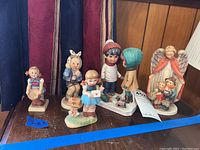 Group of five figurines on a wooden surface, including three Goebel Hummel figurines with children and two additional figurines including a Gorham angel.